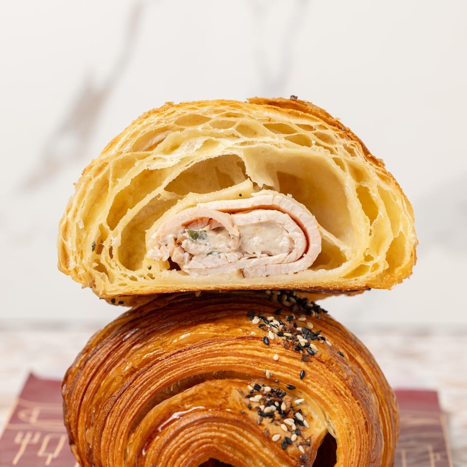 Two stacked laminated pastries, including a sliced savory chicken-filled croissant atop a sesame-seeded pain au chocolat.