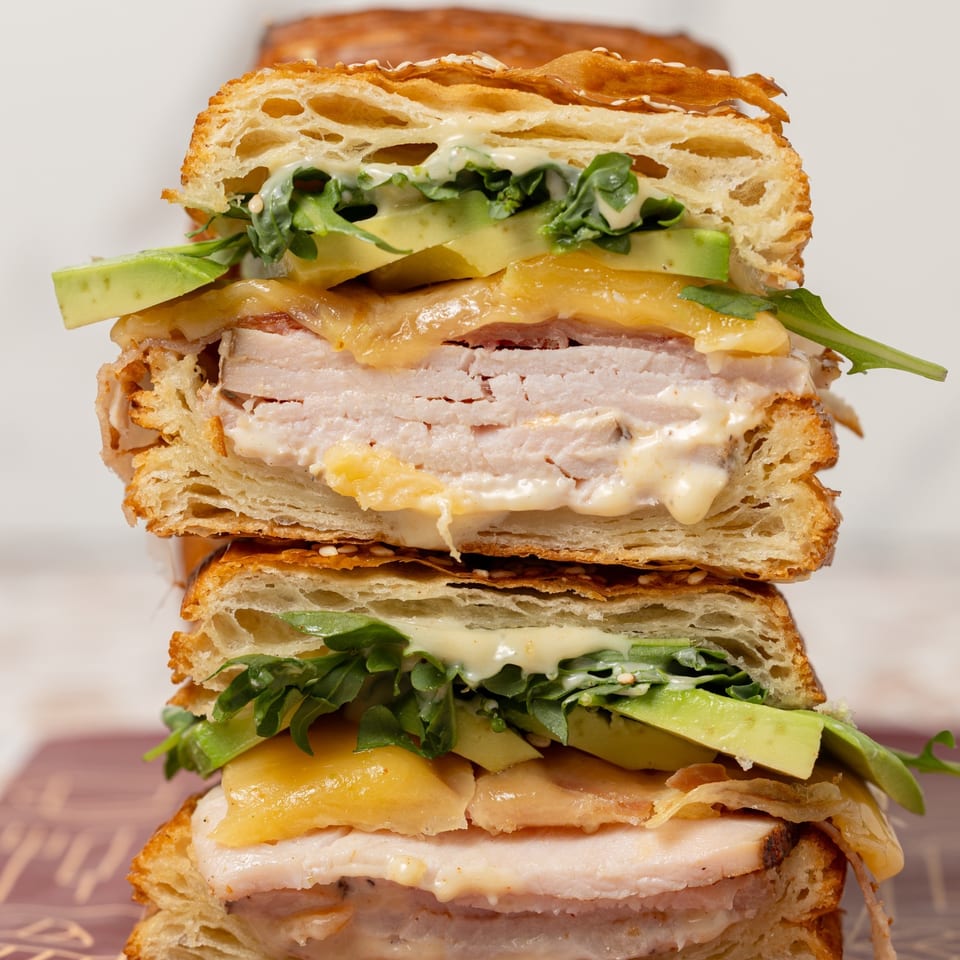 Stacked toasted sandwiches layered with sliced turkey, melted cheese, avocado, leafy greens, and creamy sauce.