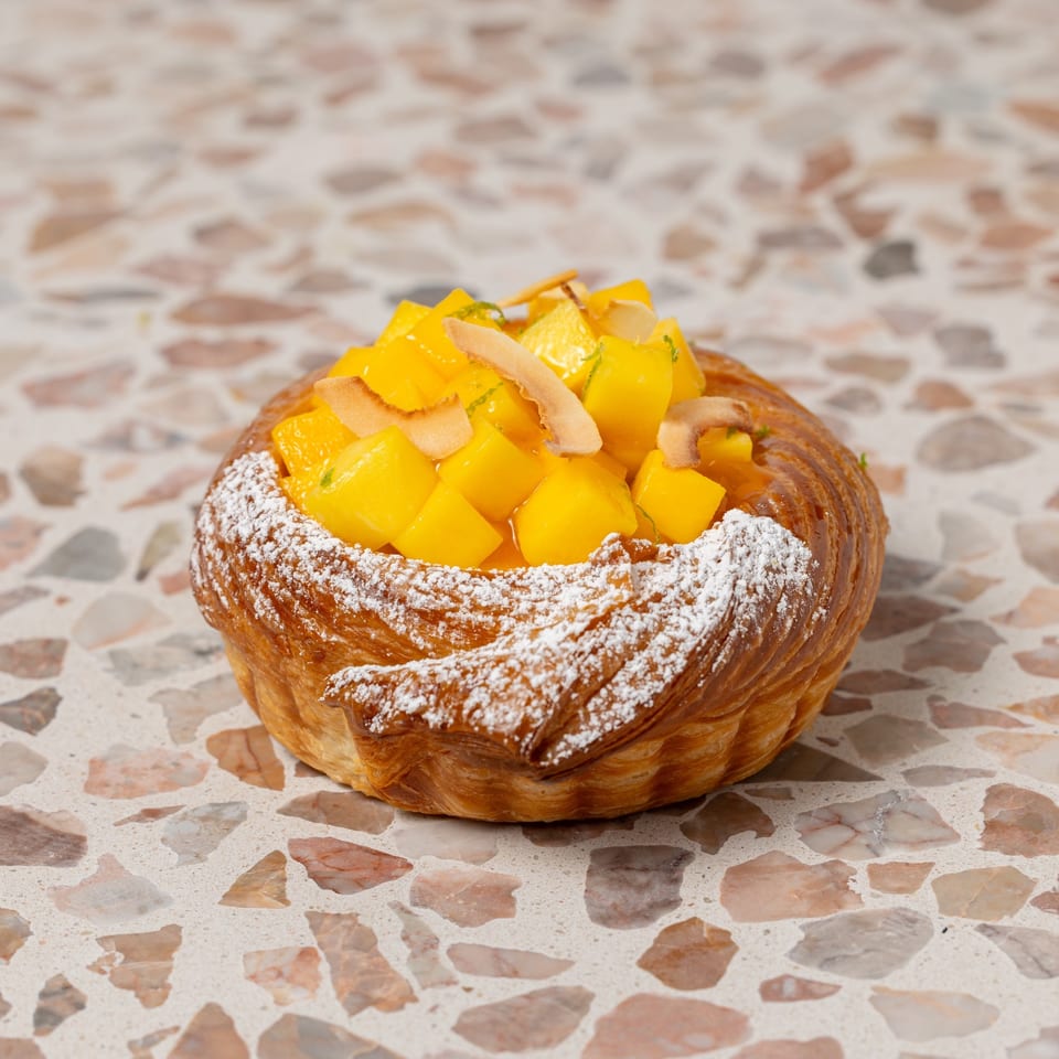 Flaky pastry topped with powdered sugar, diced mango, and coconut shavings on a terrazzo tabletop.