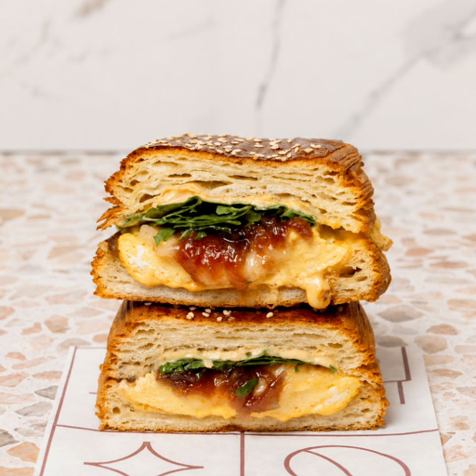 Stacked toasted sandwiches with melted cheese, greens, and reddish filling on patterned paper over a terrazzo table.