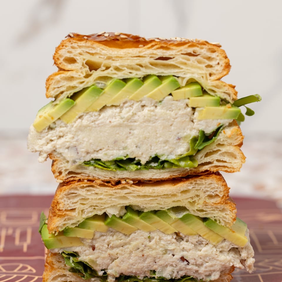 Stacked croissant sandwiches filled with chicken salad, sliced avocado, and leafy greens on a decorative tabletop.