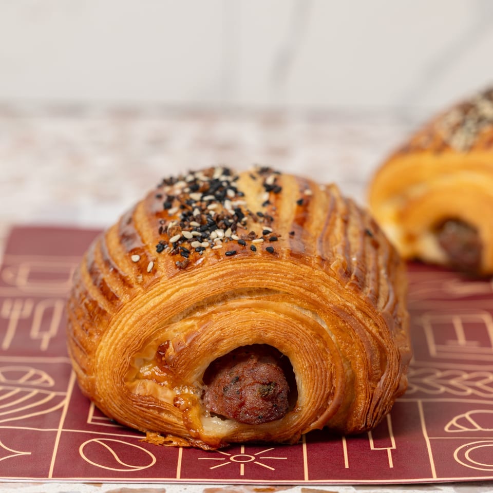 Golden laminated pastry filled with sausage and topped with sesame and poppy seeds, displayed on patterned paper.