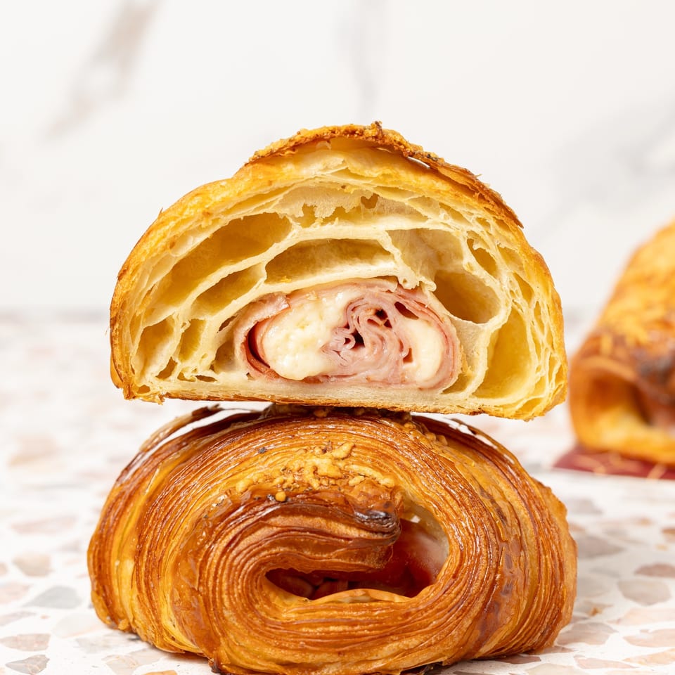 Stacked laminated pastries, including a cross-section filled with rolled ham and melted cheese, on a terrazzo surface.